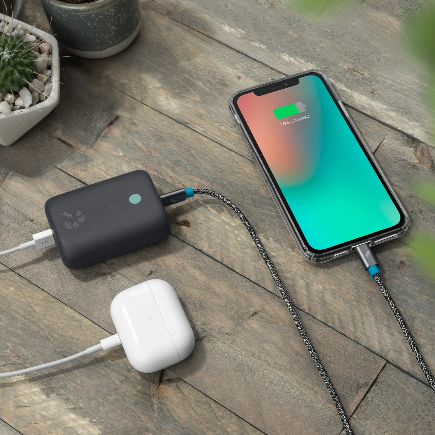 Charcoal portable charger with green button connected to airpods and cell phone on table.