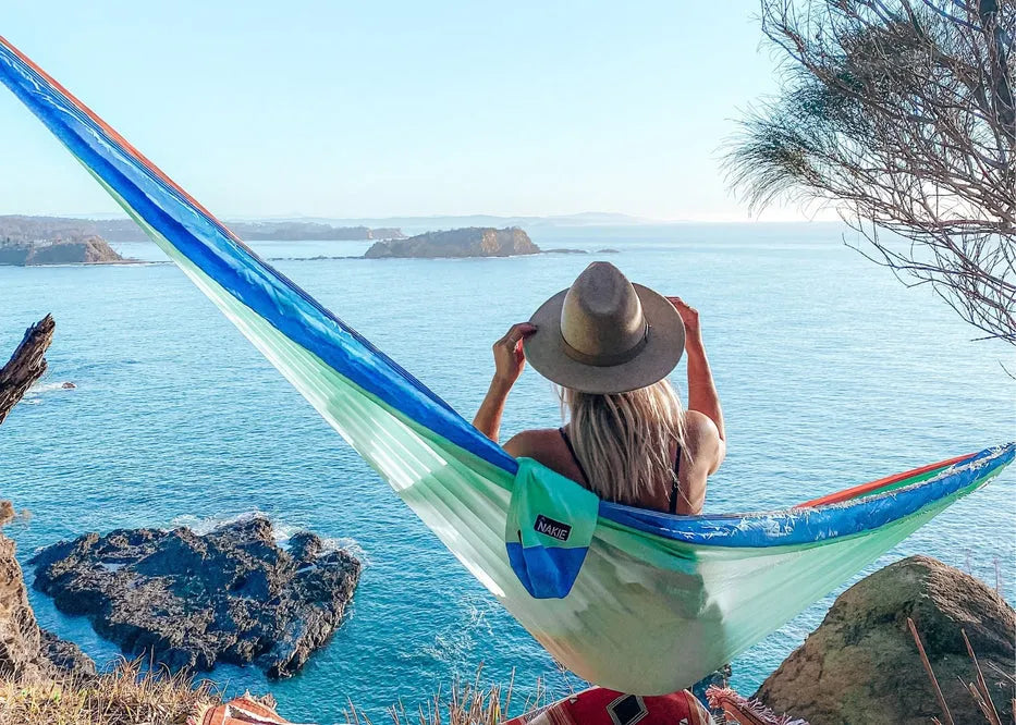 Woman sitting in a sku blue and orange hammock on a cliff overlooking water.