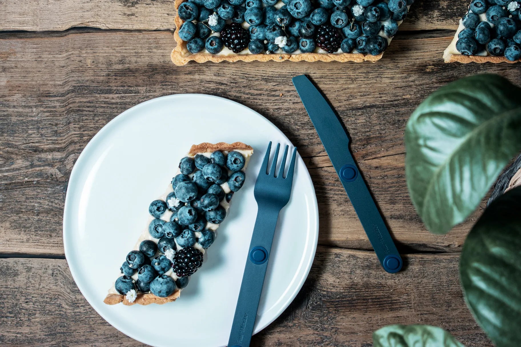 Blue fork and knife next to blueberry pie on a plate.