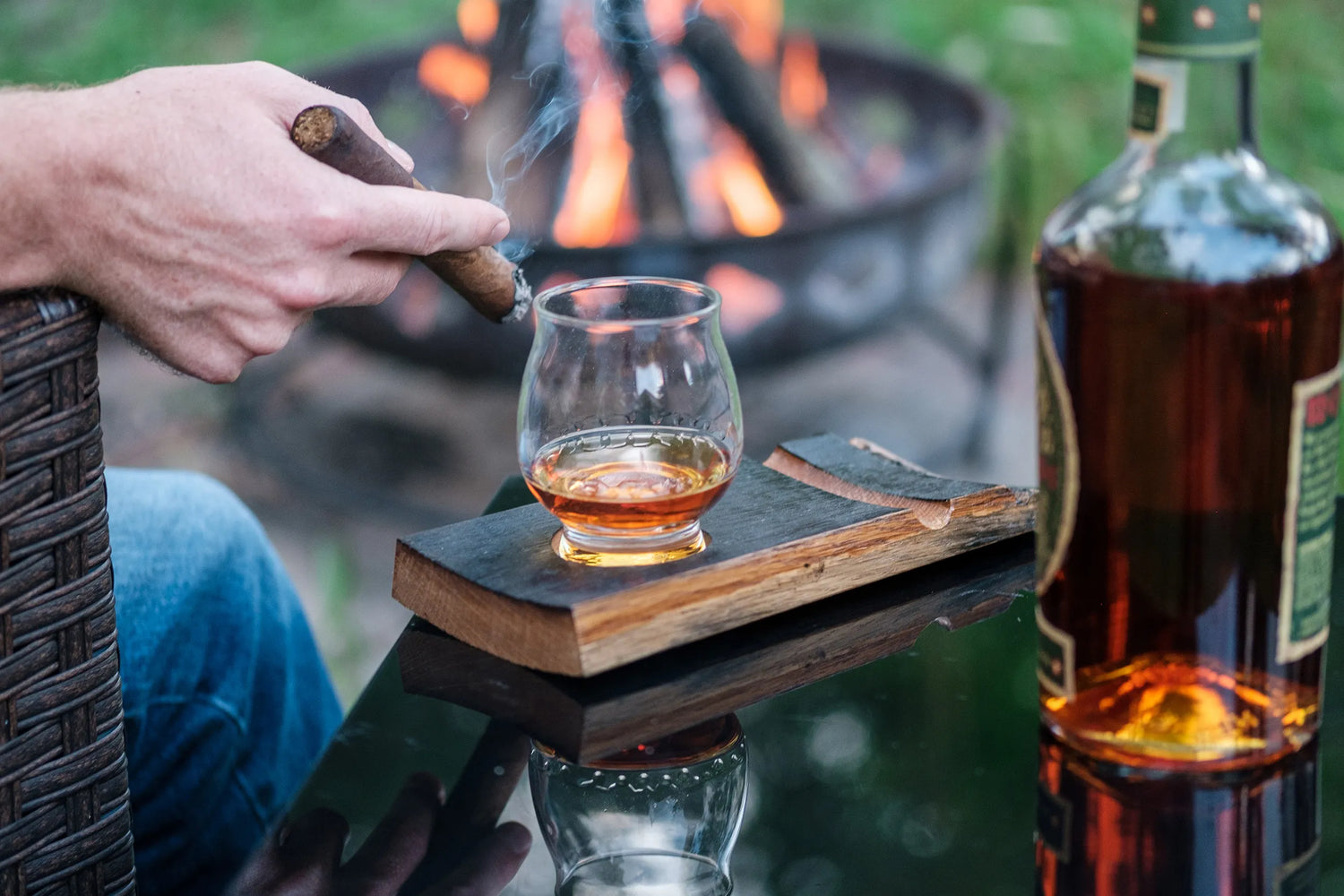 Man sitting by a fire holding a cigar next to a glass of whiskey.