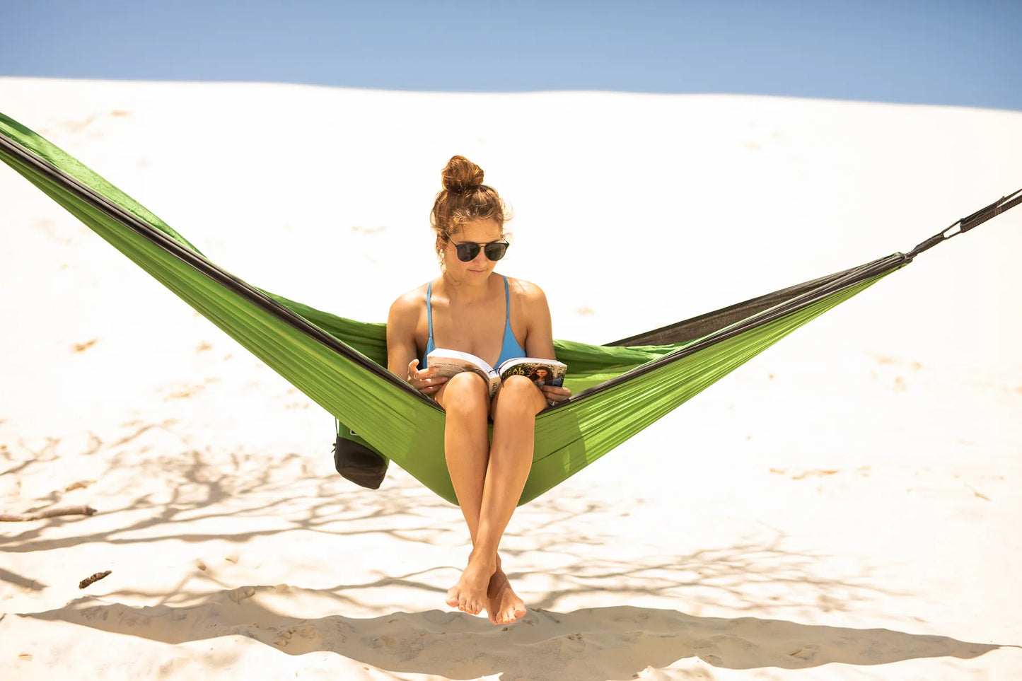 Woman on the beach sitting in a green hammock reading a book.