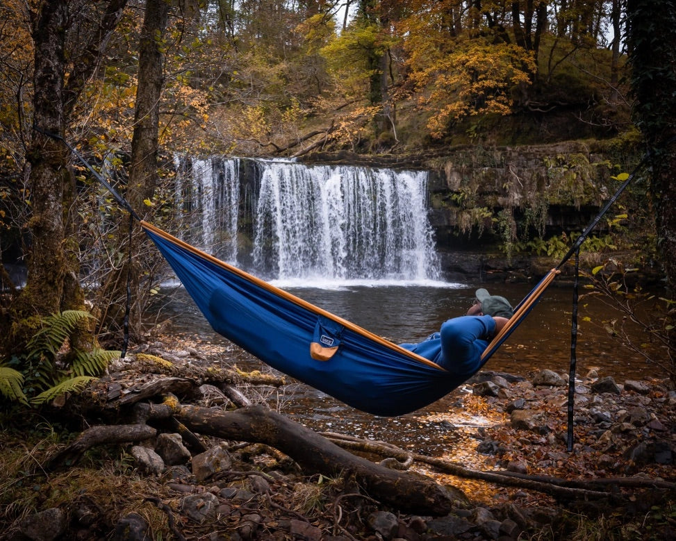 Man laying in a blue and orange hammock by a waterfall.
