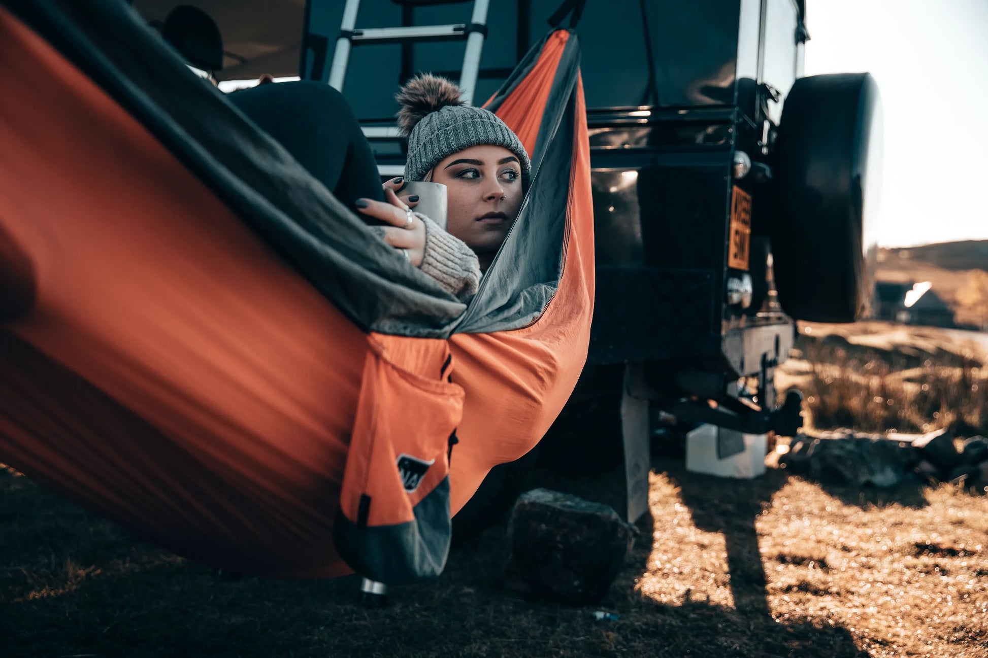 Woman lying and drinking coffee in a orange and gray hammock outside.