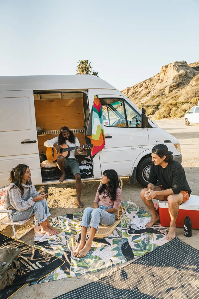 Friends sitting around on the beach in front of a van on blankets.