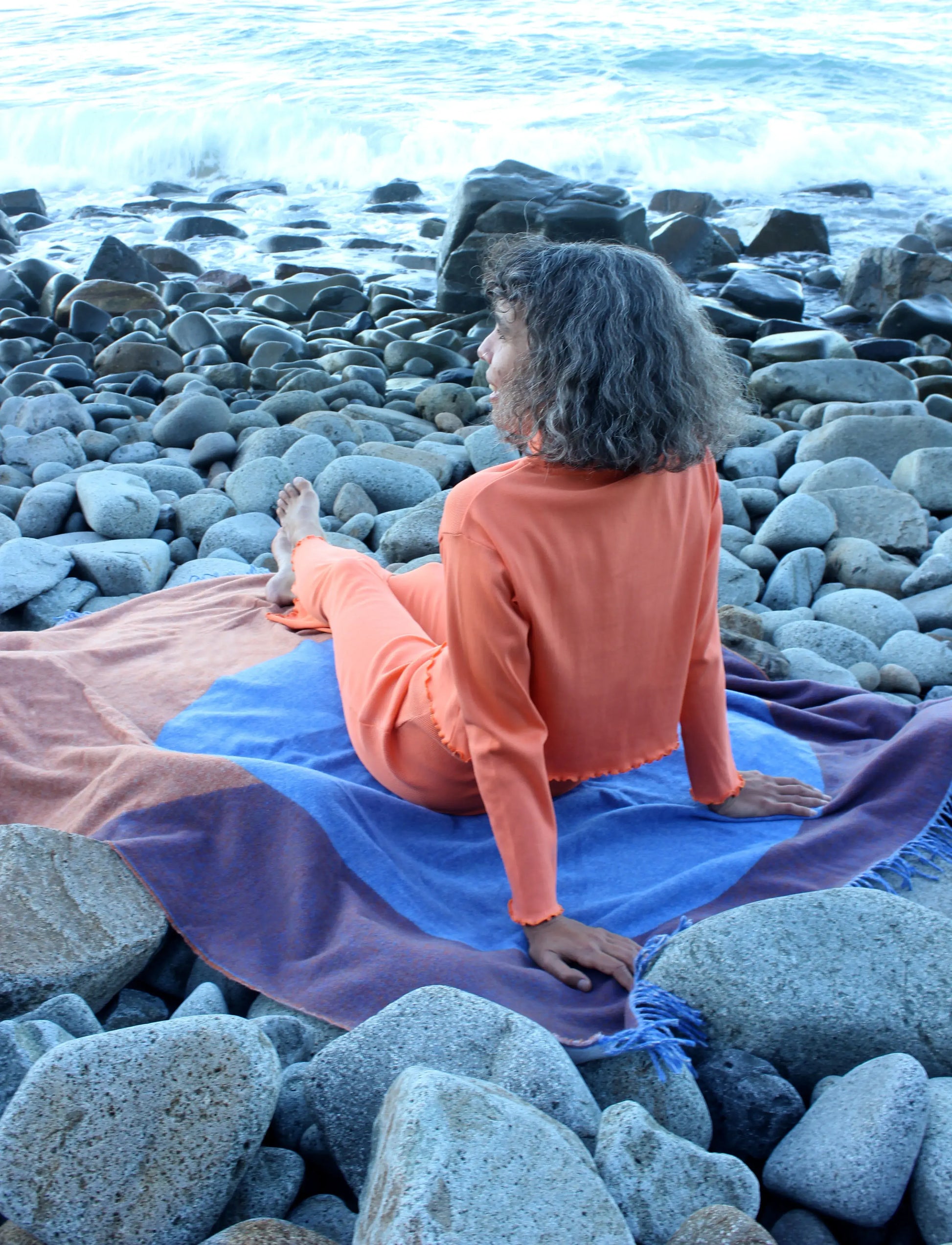 Woman sitting on a blue, orange and purple wool blanket on a rocky beach.