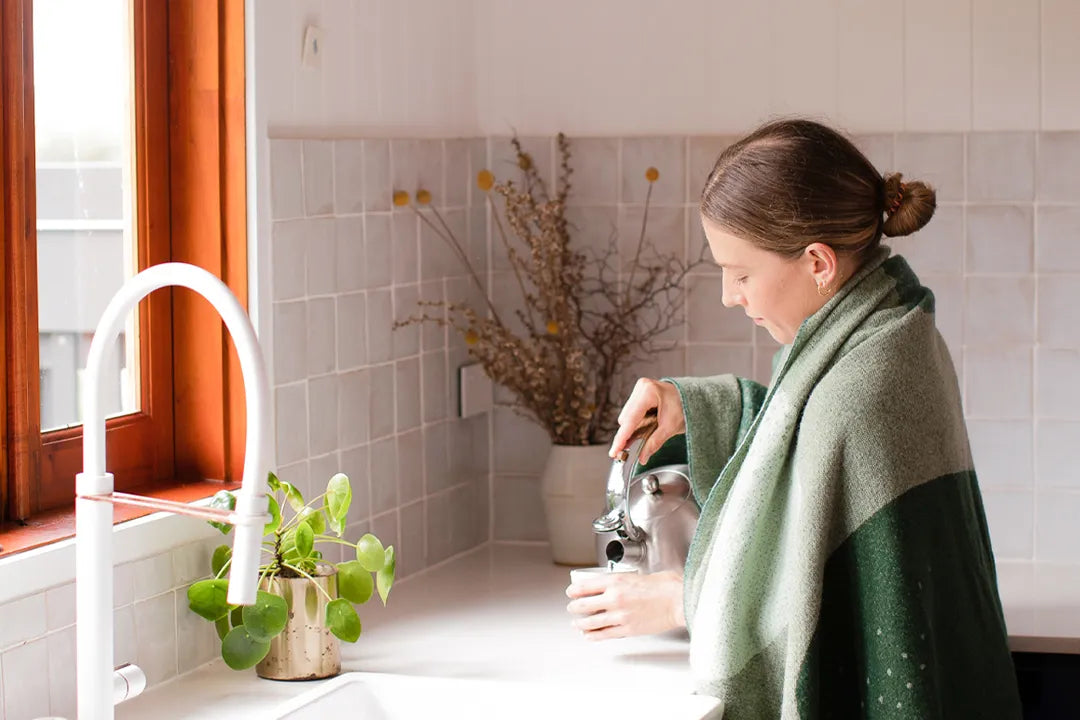 Woman in a kitchen pouring water wearing a green wool blanket.