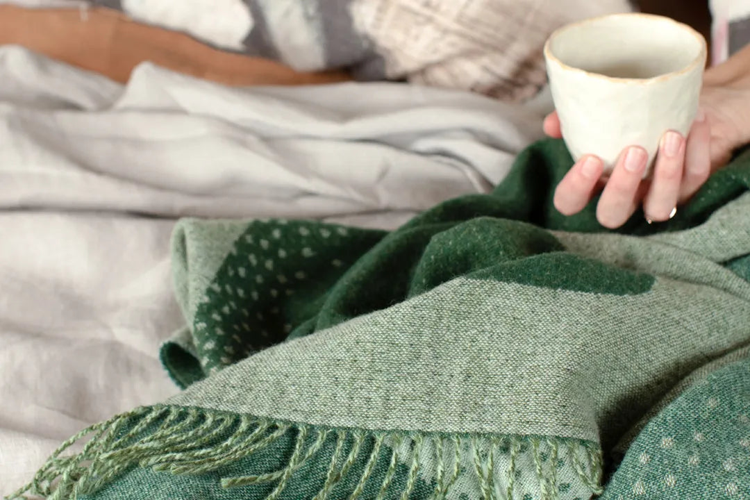 Green wool blanket on a bed with person holding a cup.
