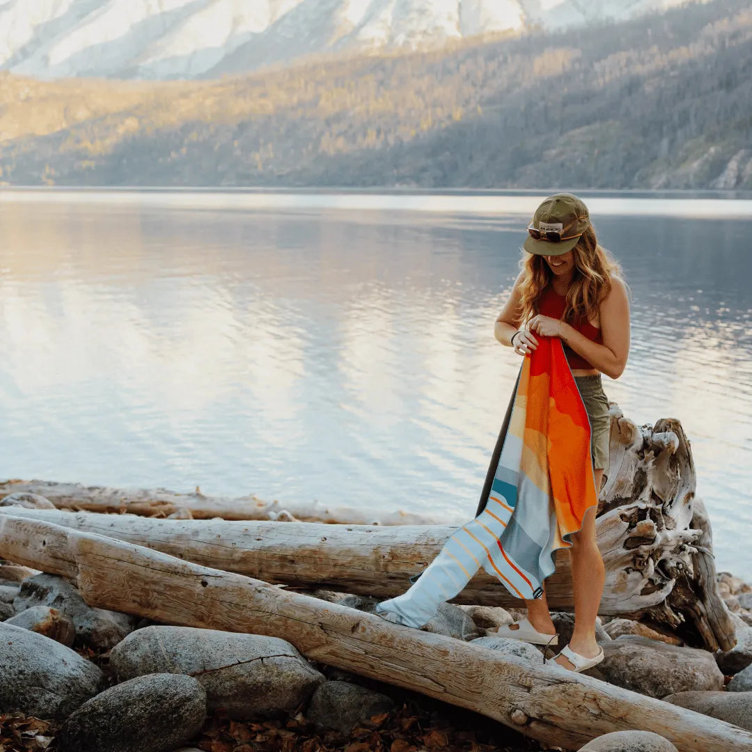 Woman outside standing next to a lake holding a multi-colored towel.