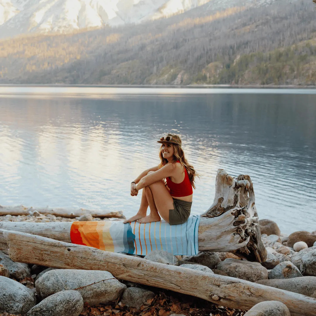 Woman outside sitting next to a lake and multi-colored towel.
