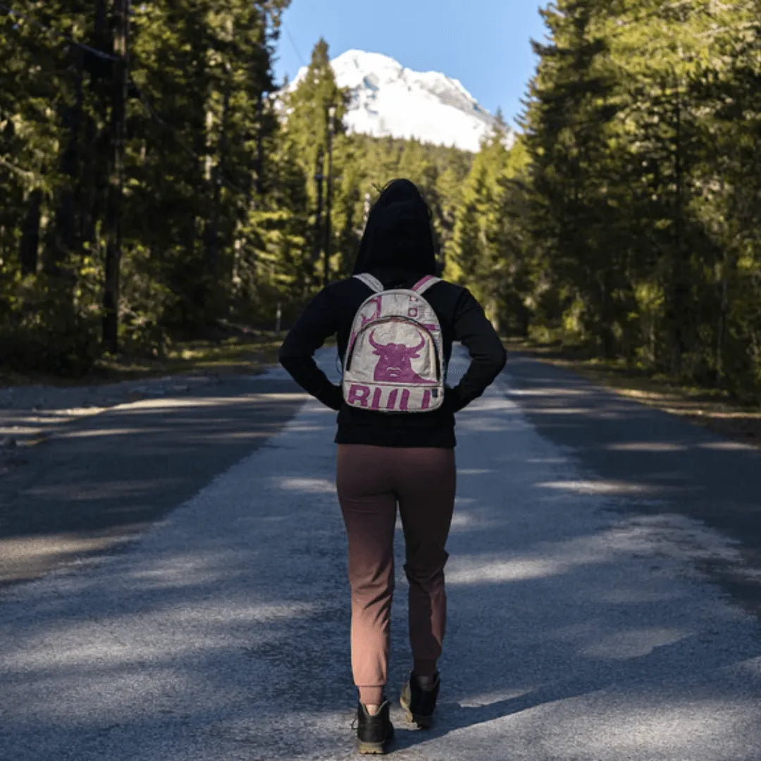 Person walking in woods wearing a white and pink backpack with a bull on it.