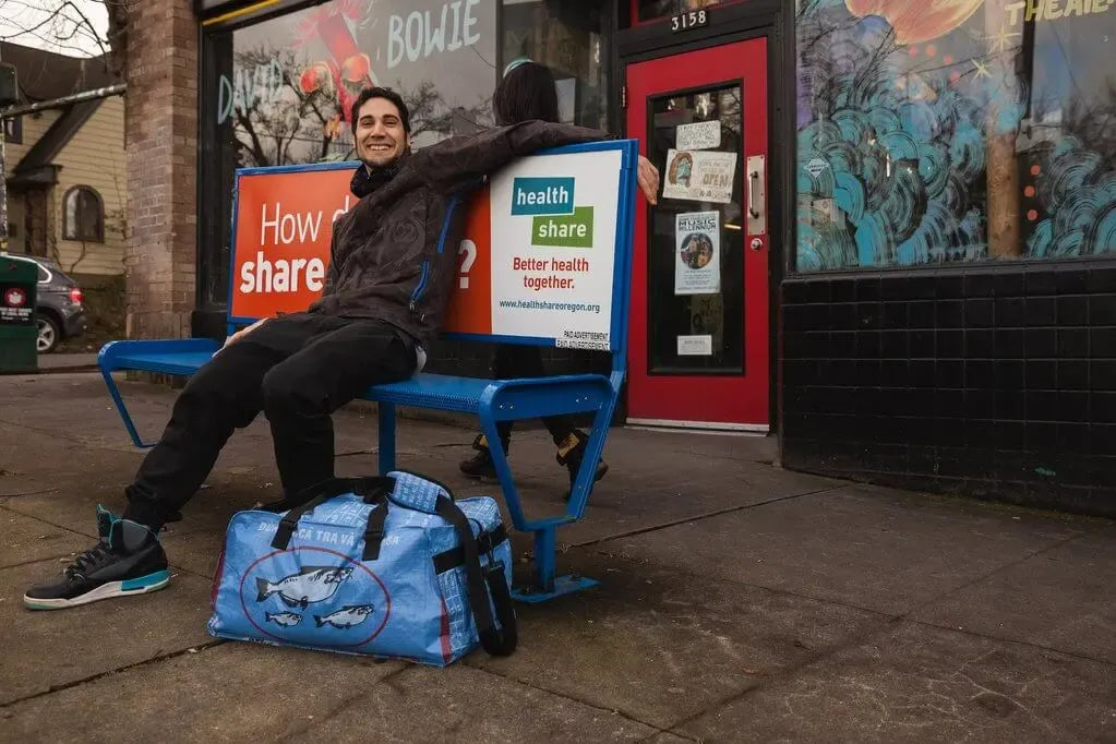 Man sitting on a bench on a sidewalk next to a blue duffel bag with fish on it.